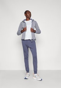 Gray patterned zip-up hoodie with drawstrings, white t-shirt, blue joggers, and gray sneakers with orange accents. Model standing with arms relaxed.