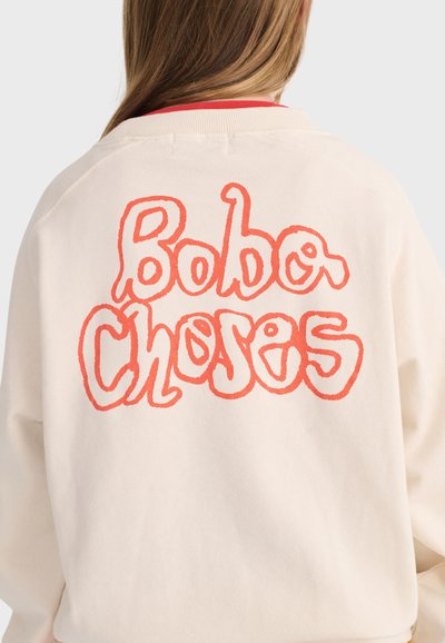 Felpa lunga color beige chiaro indossata da una persona con capelli lunghi, con grande testo arancione scritto a mano "Bobo Choses" sulla schiena.