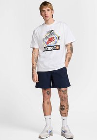 Camiseta blanca con gráficos coloridos de Nike, combinada con pantalones cortos navy. Presenta tatuajes en los brazos y zapatillas blancas con acentos azules.