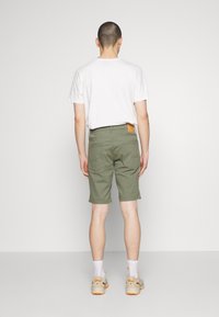 Pantalones cortos verdes con corte recto, hechos de algodón. Presentan una etiqueta de cuero en la parte trasera de la cintura y dos bolsillos traseros. Combinados con una camiseta blanca.