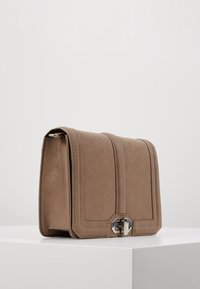 Beżowa skórzana torebka o wyraźnym kształcie, z ozdobnym przeszyciem, srebrnym zapięciem i gładką fakturą.