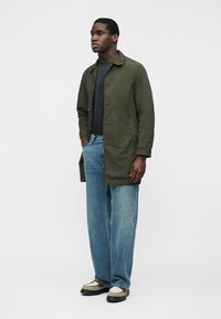 Άνδρας φορώντας παλτό ελιάς, μαύρο πουκάμισο, χαλαρά μπλε τζιν και μπεζ-καφέ παπούτσια, στέκεται με τα χέρια στις τσέπες.