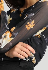 Schwarze Bluse mit floralem Muster, die durchsichtige lange Ärmel mit elastischen Bündchen hat, verziert mit gelben und grauen Blumen und einem taillierten Schnitt.