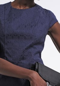 Granatowa, teksturowana sukienka z geometrycznym wzorem, z krótkimi rękawami; trzyma czarną kopertówkę o eleganckim designie i srebrnych akcentach.