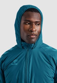 Teal zip-up hoodie med huva, som har en logotyp på bröstet och mönstrade detaljer på axlarna. Slät, lätt tyg.