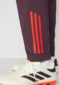Donkerpaarse sportbroek met drie verticale reflecterende oranje strepen aan de zijkant, gecombineerd met witte voetbalschoenen met zwarte accenten.