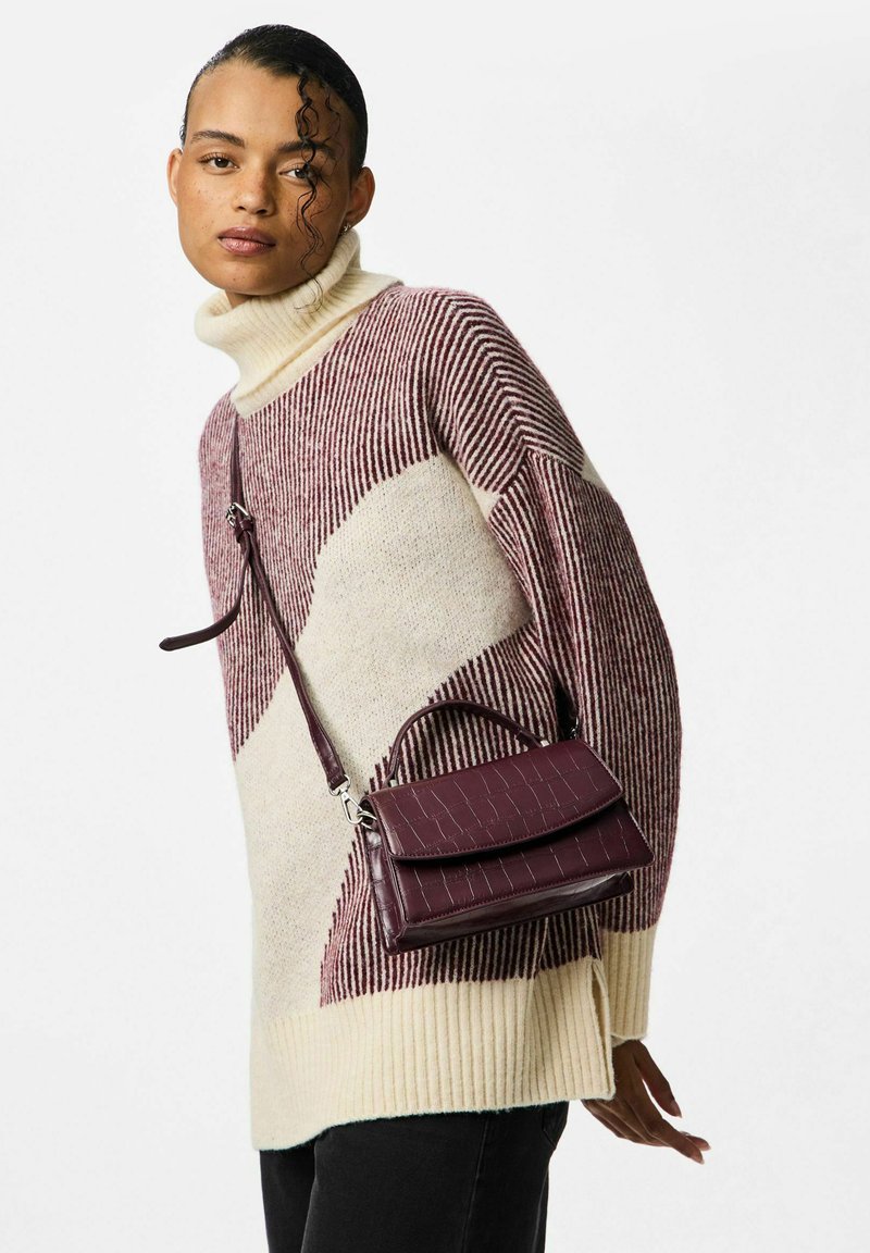 Kvindelig iført en cremet og burgunderstribet turtleneck sweater og sorte bukser, bærende på en burgunder læder crossbody taske.