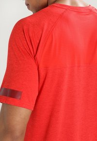 Czerwona sportowa koszulka z krótkimi rękawami, teksturowana tkanina i siateczkowy panel na górnej części pleców. Posiada odblaskowy akcent na rękawie.