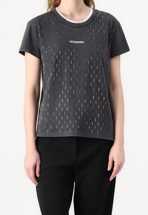 Žena s černým tričkem s krátkým rukávem s perforovaným vzorem a logem "PATRIZIA PEPE" na hrudi, v kombinaci s černými kalhotami.