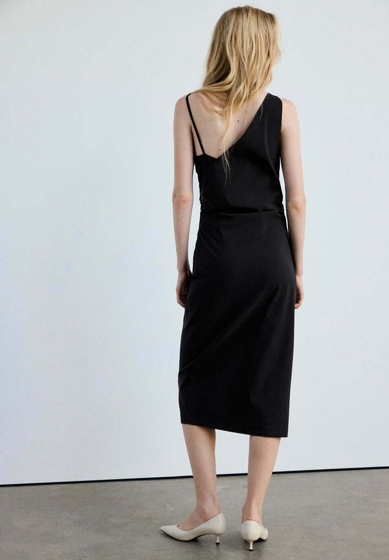 Femme aux longs cheveux blonds portant une robe midi asymétrique noire et des chaussures beige à talons petits, debout contre un mur uni.