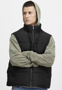 Schwarze Steppweste mit hohem Kragen, Reißverschluss und ärmellosem Design. ÜBer einem hellgrünen langärmeligen Hoodie getragen. Lässige Jeans.