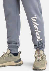 Graue Jogginghose mit einem weißen "Timberland"-Logo und Baumemblem am linken Bein. Sie haben eine weiche Textur und Bündchen an den Knöcheln. Tragen Sie sie mit beigen Turnschuhen.