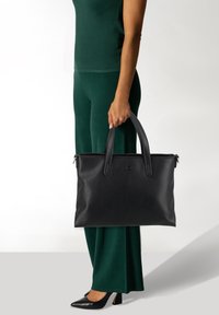 Borsa tote nera in pelle con doppi manici; design elegante, texture liscia e chiusura con zip. Abbinata a pantaloni verdi e tacchi neri.