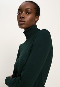 Frau mit geflochtenem Haar, die einen dunkelgrünen, gerippten Rollkragenpullover trägt und vor einem hellen, einfarbigen Hintergrund posiert.