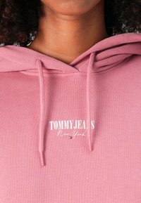 Un sweat à capuche rose avec une poche kangourou à l'avant, un cordon de serrage au niveau de la capuche et un logo blanc "TOMMYJEANS New York". Tissu doux avec des détails de couture texturés.
