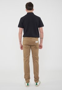 Polo shirt nero, pantaloni beige e sneakers con accenti verdi. I pantaloni presentano tasche posteriori e un dettaglio con etichetta, design semplice con una forma aderente.