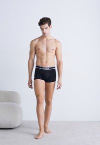 Boxer-briefs noirs avec une ceinture logotée blanche "LACOSTE", présentés sur un modèle au physique tonique, se tenant sur une surface de couleur claire.