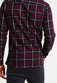 Uomo che indossa una camicia a maniche lunghe aderente rossa e blu a quadri abbinata a pantaloni neri, mostrato di spalle su uno sfondo neutro.