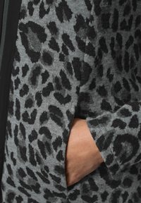 Mano parzialmente dentro della tasca di un giubbotto di stoffa grigia con stampa leopardata nera e zip nera visibile.