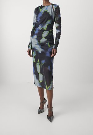 Mannequin portant une robe midi à manches longues froncée avec un motif abstrait bleu, noir et vert, associée à des escarpins métalliques à bout pointu.