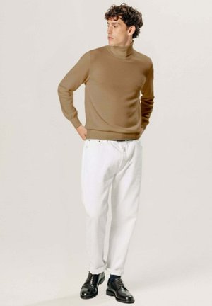 Mann, der einen beige Rollkragenpullover, weiße Hosen und schwarze Anzugschuhe trägt, steht mit den Händen hinter dem Rücken und schaut zur Seite.