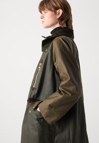 Olijfgroene, waterdichte jas met een rits aan de voorkant, corduroy kraag en gouden accenten. Heeft zijzakken en een relaxte pasvorm.
