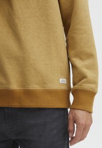 Sudadera marrón con un tejido texturizado y aterciopelado, con puños y dobladillo acanalados, además de una pequeña etiqueta en el lado inferior.