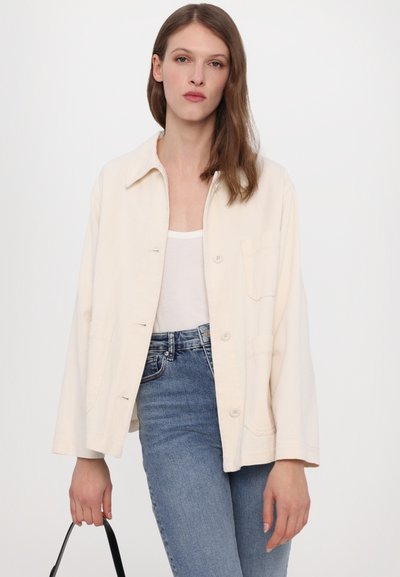 Valkoinen overshirt, jossa on kauluksellinen kaulus, etunapit ja läppätaskut, yhdistettynä vaaleansinisiin farkkuihin ja mustaan laukkuun.