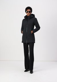 Zwarte geïsoleerde parka met een capuchon, voorrits en twee zijzakken. Gladde textuur, slim fit, gedragen met zwarte broek en schoenen.