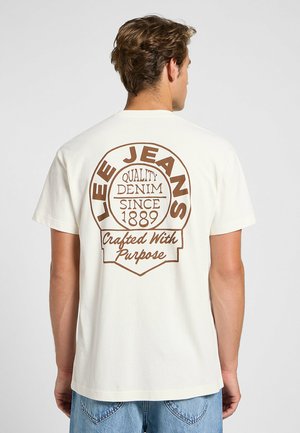 Camiseta crema con un gran gráfico marrón que incluye el texto "LEE JEANS", "QUALITY DENIM" y "Crafted With Purpose". Mangas cortas.