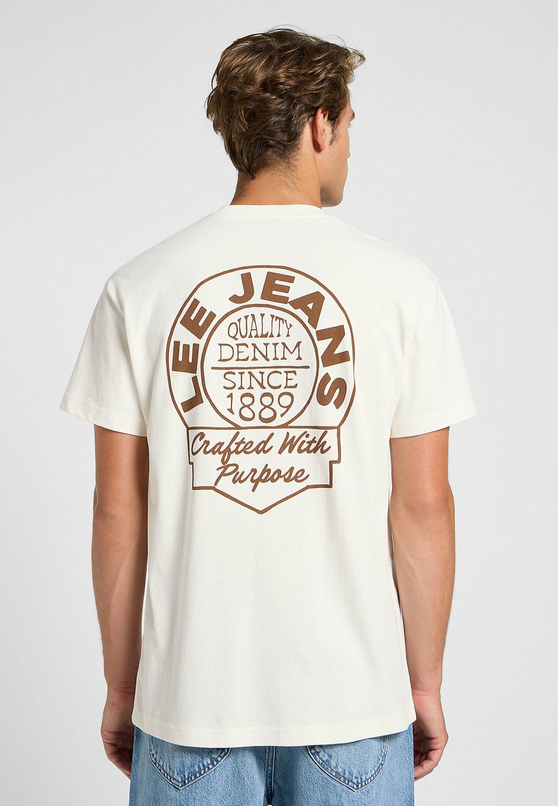Crème t-shirt met een grote bruine grafische afbeelding met tekst, waaronder "LEE JEANS," "QUALITY DENIM," en "Crafted With Purpose." Korte mouwen.