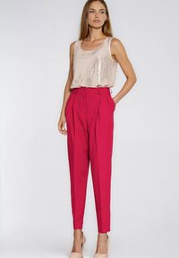 Top na ramiączkach z cekinami w jasnoróżowym kolorze, zestawiony z wysokim stanem i plisowanymi spodniami w jaskrawym różu. Modelka nosi buty na obcasie w neutralnym odcieniu.