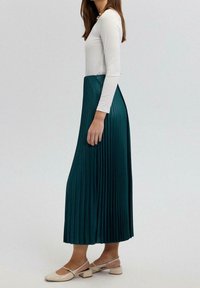 Plissé, teal maxi rok met een hoge taille en vloeiende stijl. Draagt een aansluitend wit langesleef top en beige blokhak schoenen.
