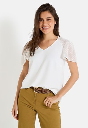 T-shirt blanc à col V avec des manches bouffantes transparentes texturées, porté avec un pantalon beige et une ceinture en imprimé léopard marron. Design sobre, tissu doux.