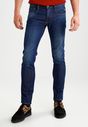 Man draagt strak zittende blauwe spijkerbroek met vervaagde details en zwarte schoenen met luipaardprint accent, staand tegen een witte achtergrond.