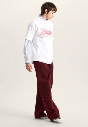 Biała koszulka z czerwonym nadrukiem na warstwie niebiesko-białej koszuli w paski, zestawiona z szerokimi czerwonymi spodniami i butami w wzory.