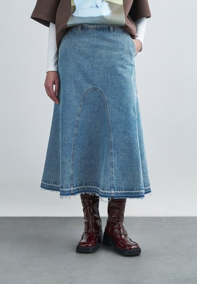 Jupe trapèze en denim bleu clair avec des détails de coutures courbes et un ourlet effiloché. Associée à des bottes montantes bordeaux brillantes.