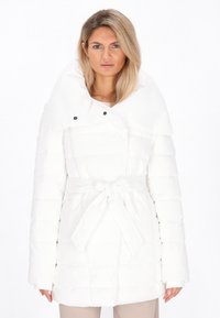 Blond kvinde iført en hvid belagt puffjakke med bred krave, der står mod en ensfarvet hvid baggrund.