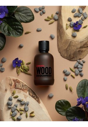 Bouteille en verre marron avec un capuchon noir, étiquetée "Original Wood DSQUARED2" en texte noir gras. Entourée de feuilles vertes, de violettes et de pierres.