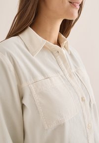 Camicia button-up beige chiaro realizzata in tessuto testurizzato. Presenta un colletto classico, due tasche frontali e dettagli di cucitura a vista.