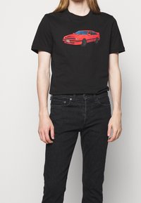 Camiseta de algodón negra con un gráfico de un coche rojo. El modelo lleva pantalones ajustados negros. Diseño minimalista con un escote redondo.