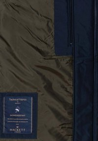 Duschbeständige Jacke mit einem dunkeloliven Außenmaterial, einem marineblauen Akzent und einem Etikett mit Angaben zu den Stoffeigenschaften. Glatte Textur und Reißverschluss.