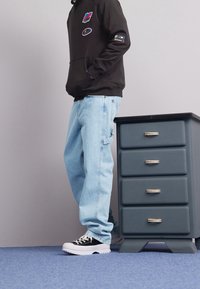 Personne portant un sweat à capuche noir et un jean bleu clair se tient à côté d'un meuble gris à quatre tiroirs avec des poignées argentées sur un tapis bleu.