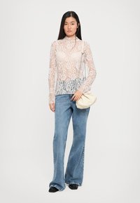 Kanten lange mouw top met bloemenpatroon in roomkleur, gecombineerd met lichtblauwe flared jeans. Het model houdt een roomkleurige clutch in haar hand.