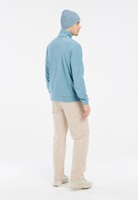 Šviesiai mėlynas fliso megztinis su aukštu apykakle, derinamas su bežiniais cargo džinsais ir derančiu kepurėle. Tekstūruota medžiaga ir atsitiktinis dizainas.