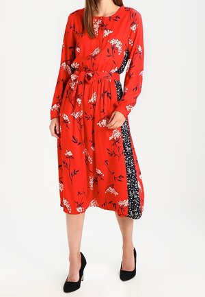 Robe midi rouge à imprimé floral blanc, manches longues, ceinture à la taille et panneaux latéraux noirs à motifs, portée avec des talons hauts noirs.