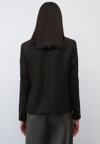 Blazer noir ajusté avec des épaules structurées, une fente unique à l'arrière et des détails de poignets à boutons. Tissu lisse avec une silhouette élancée.