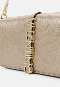 Bolso metálico dorado con un acabado texturizado, que presenta un acento de logo dorado y una correa negra en contraste. Diseño rectangular compacto.