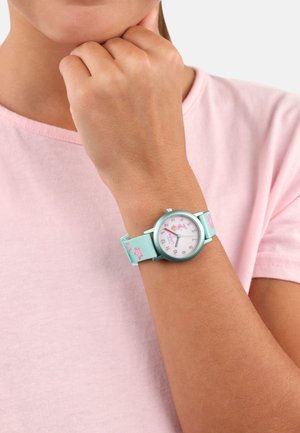Eine Kinderuhr mit einer runden silbernen Lünette, einem rosa Zifferblatt und einem blauen Stoffarmband mit rosa Sternmustern. Am Handgelenk getragen.