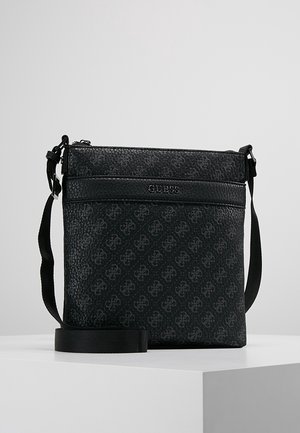 Sac bandoulière noir Guess avec sangle ajustable, motif texturé et fermeture éclair sur le dessus, posé sur une surface blanche avec un fond gris.